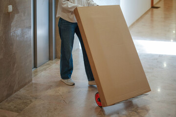 Woman pushes cart with large cardboard box in modern hallway. Online order, delivery, moving to new home concept