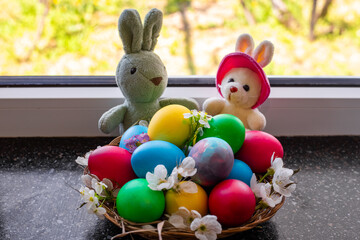A basket filled with brightly colored Easter eggs and white flowers, flanked by two stuffed bunnies on a windowsill. Easter.