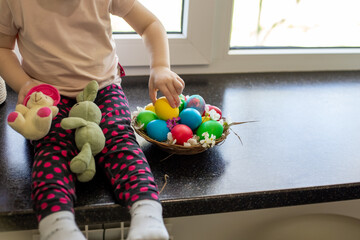 A child sits on a tabletop, happily selecting a yellow Easter egg from a basket of colorful eggs surrounded by stuffed bunnies.