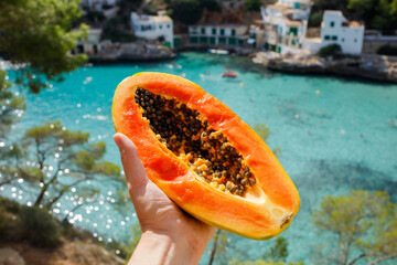 Half of fresh ripe papaya cut open with seeds in woman hand with the background of shining turquoise sea bay in summer sunny day