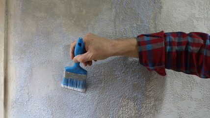 Hand of a man in a plaid shirt holds a blue brush and applies liquid primer on a textured plastered gray wall, close-up, priming a home wall with uneven textured plaster using a brush