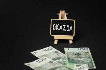 Black wooden writing board on a black background with the inscription "Okazja", in the background scattered Polish złoty PLN banknotes (selective focus)