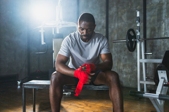 Fitness workout in gym. African American man fighter preparing for fight wrapping hands with red boxing wraps sports protective bandages in gym. Strong man ready for fight boxing sparring training - Powered by Adobe