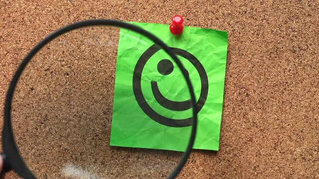 A person with a magnifying glass in his hand magnifying a green note paper with a happy face on it. Close up.
