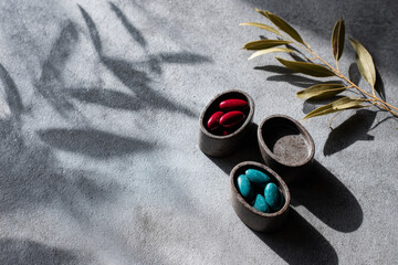 Colorful stones in oval containers, red and blue hues, placed on textured gray surface with soft shadows and green leaves, creating a serene and artistic atmosphere
