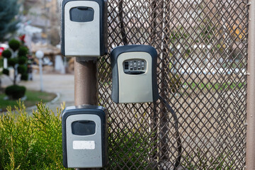 three grey and black key lockboxes mounted on a metal fence post against a background of wire mesh and blurred greenery.