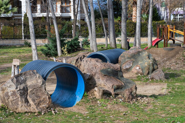 a playground obstacle course featuring blue corrugated tunnels embedded in artificial rock formations, set within a grassy park lined with tall, thin trees.