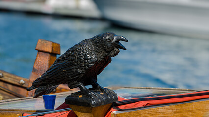 Close-up of a carved black bird on a wooden ship, with out of focus boats in the background.