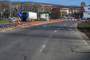 a road construction zone featuring orange cones, a white semi-truck, and traffic signs directing vehicles to the left lane.