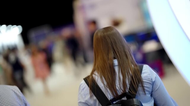 Rear view of an attendee with long hair and backpack moving through a busy indoor exhibition hall, with shallow depth of field highlighting blurred booths, networking crowds, and bright event lighting