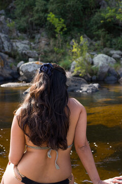 Mujer con canas de espaldas usando bikini esta descansando sobre una roca al lado del rio