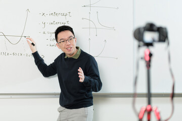 A Chinese mathematics instructor wearing glasses teaches concepts on a whiteboard while being recorded for an online lesson, ideal for online education, math tutoring, or STEM education concepts.