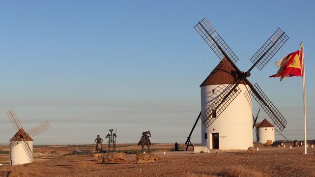 Famous windmills of La Mancha with Don Quixote, Sancho Panza statues and Spanish flag waving at sunset
