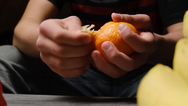 Hands of a young man peel a mandarin. A person removes mandarin peel with fingers. Peeling an orange by hands. Citrus fruits are peeled manually. Natural vitamins are contained in fruits.