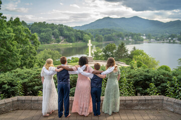 Five children stand on a stone overlook with arms around each other, viewing a scenic lake and distant mountains with a white cross.