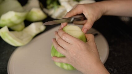 Unidentifiable woman peeling a whole winter melon with a knife on a cutting board. She then cuts the wax gourd into large pieces for cooking