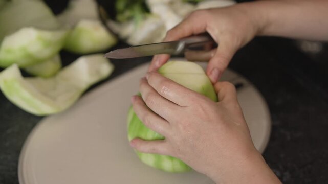 Unidentifiable woman peeling a whole winter melon with a knife on a cutting board. She then cuts the wax gourd into large pieces for cooking
