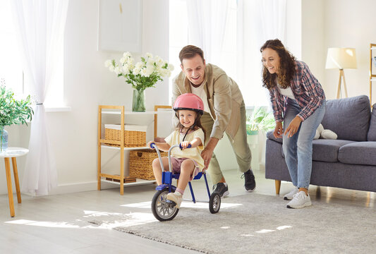 Happy family parents and daughter play at home riding bicycle. In livingroom, child girl rides tricycle in helmet as dad guides and mom cheers. Fun, safety, supportive parenting and leisure concept.