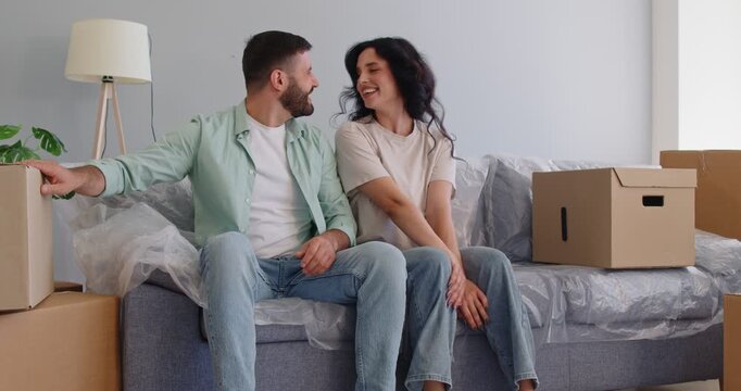 Happy couple sitting on sofa by moving boxes and lamp to enjoy romantic first day in new home with laughter. Young smiling man and woman hugging with love and care in living room full of cartons
