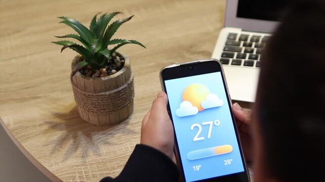 View from behind a young individual holding a cell phone and looking at the weather forecast app, with a laptop and a potted plant on the wooden table in the distance