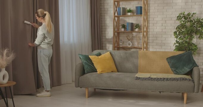 A woman uses a lint roller to clean curtains in a stylish living room with a cozy grey sofa and colorful pillows.