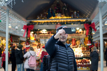 Adult male capturing memories with a mobile phone at a festive holiday fair with blurred lights and decorations in the background.
