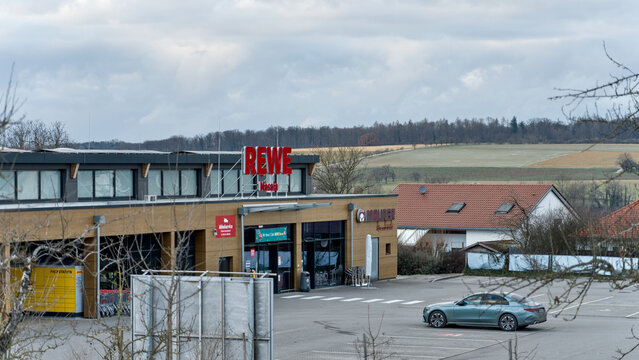 REWE supermarket with a parking lot and a car, set against a rural landscape in the background.