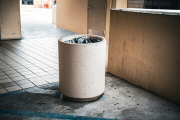 Concrete trash can with black liner sits on a tiled walkway next to a wall