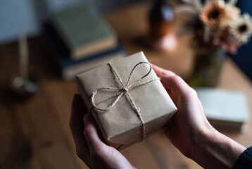 Hands hold a small brown paper-wrapped gift tied with string. On a wooden table, there are books and a vase of flowers nearby. The scene shows a cozy indoor setting