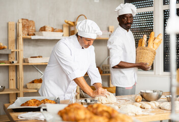 Skilled middle-aged baker in white uniform kneading dough on flour-dusted wooden table in cozy inviting setting of small artisan bakery, highlighting craftsmanship of traditional breadmaking..