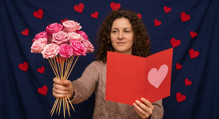 A woman displays a handmade bouquet of pink paper roses and a red greeting card with a heart against a dark blue background decorated with small hearts.