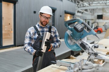 A carpenter cuts a wooden plank on a sawmill in a modular building factory, providing precise cuts for a prefabricated structure