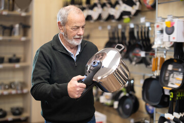 Senior man customer inspect product range and selects stewpot. Client in hardware store picks up household goods.