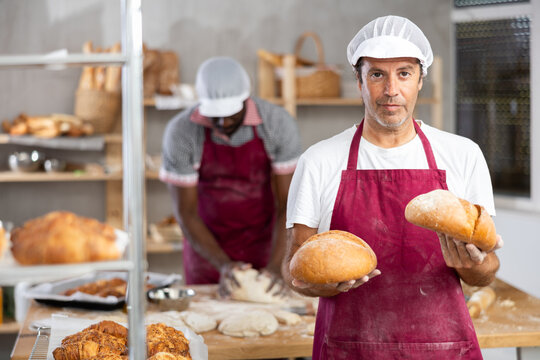Caucasian man baker with loafs of bread, shows many products. He has made large number of fresh fragrant baguettes, filled basket with pastries and ready to provide customers with fresh products goods