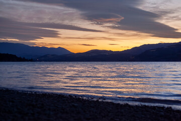 Obraz premium Tranquil sunset over alpine lake with mountain silhouettes and golden reflections. Peaceful evening scene in European mountain region.