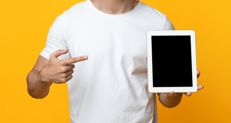 Unrecognizable guy in white t-shirt pointing at pad, cropped of man showing digital tablet with blank screen, mockup, yellow studio background, panorama, closeup. Mobile application concept
