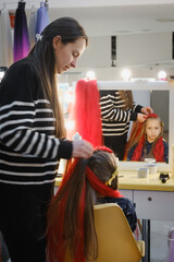 Woman braider working on young girl's red synthetic braids in beauty salon retail setting, showcasing professional hair styling service with product displays and modern store fixtures.