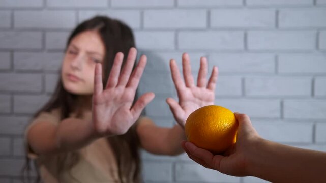 Citrus allergy. Young girl showing a strong allergic reaction or intolerance to citrus fruit, rejecting an orange offered by a hand with a determined stop gesture, symbolizing food allergies