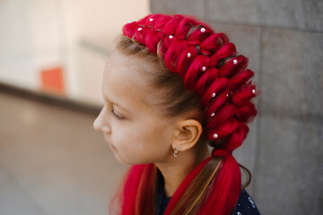  Portrait of smiling 7 years old girl with eye-catching red synthetic braids arranged in decorative twin braided buns with pearl accents, navy patterned top, casual indoor setting on gray wall
