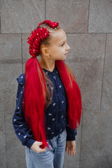 Young girl with elaborate red braided updo hairstyle featuring synthetic hair extensions decorated with pearl beads, wearing shirt and denim, against gray tiled wall