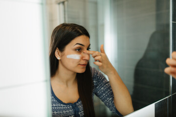 Young female in a white bathrobe applying facial moisturizer while looking in the bathroom mirror