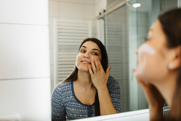 Young female in a white bathrobe applying facial moisturizer while looking in the bathroom mirror