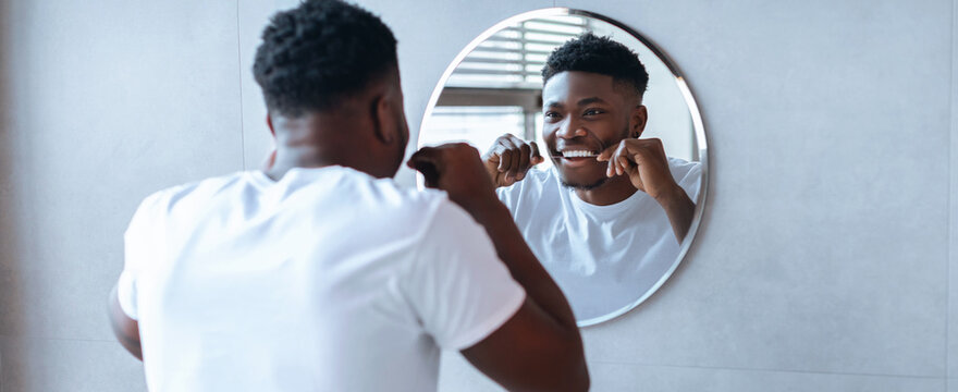 A man stands in front of a round mirror in a bathroom. He is brushing his teeth while looking at his reflection. The room is well-lit and has a modern design.