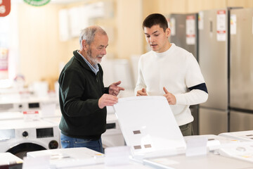 Eldery and young man buyers chooses washing machine or dishwasher in home appliance store..