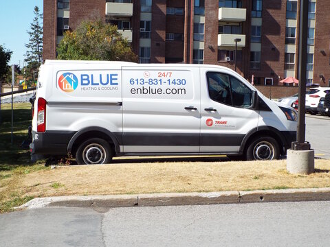 Gloucester, ON, Canada-July 3,2025:A white "Blue Heating and Cooling" service van is parked in a lot, featuring company contact information and a Trane logo against a brick apartment on Innes Rd.
