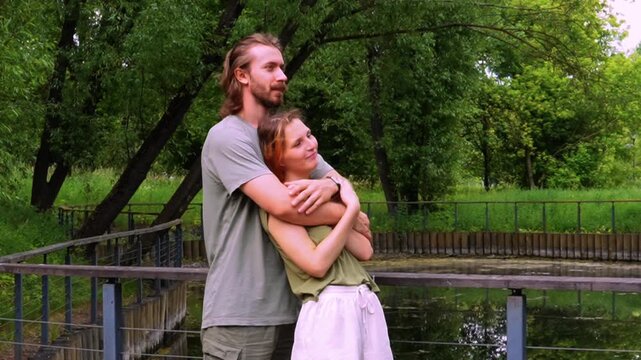 tender embrace of young loving couple outdoors in lush green summer forest man with long hair beard holding woman with reddish hair as she leans head on his chest both smiling gently eyes closed