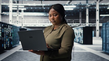 Technician in server room using laptop to upgrade rigs for increase in clients workload capacity. African american woman in data center configures equipment, improving processing speed, camera A