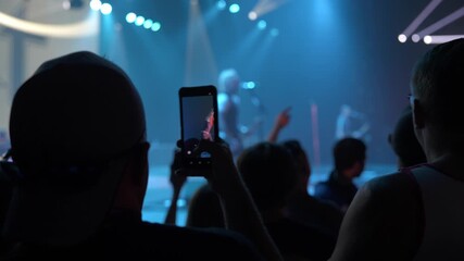 Silhouetted fans watch a live concert as a smartphone records the stage under blue lights. Raised hands and spotlights create a loud festival atmosphere inside the venue.