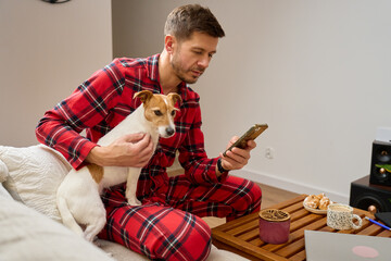Man sitting on couch in pajamas holding dog and using smartphone