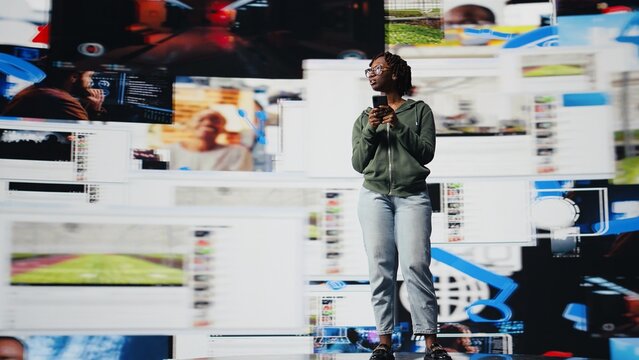 Happy woman uses smartphone to do texting with friends, discussing movies, standing in front of vibrant video wall collage. Teenager chatting with other social media users using mobile phone, camera A